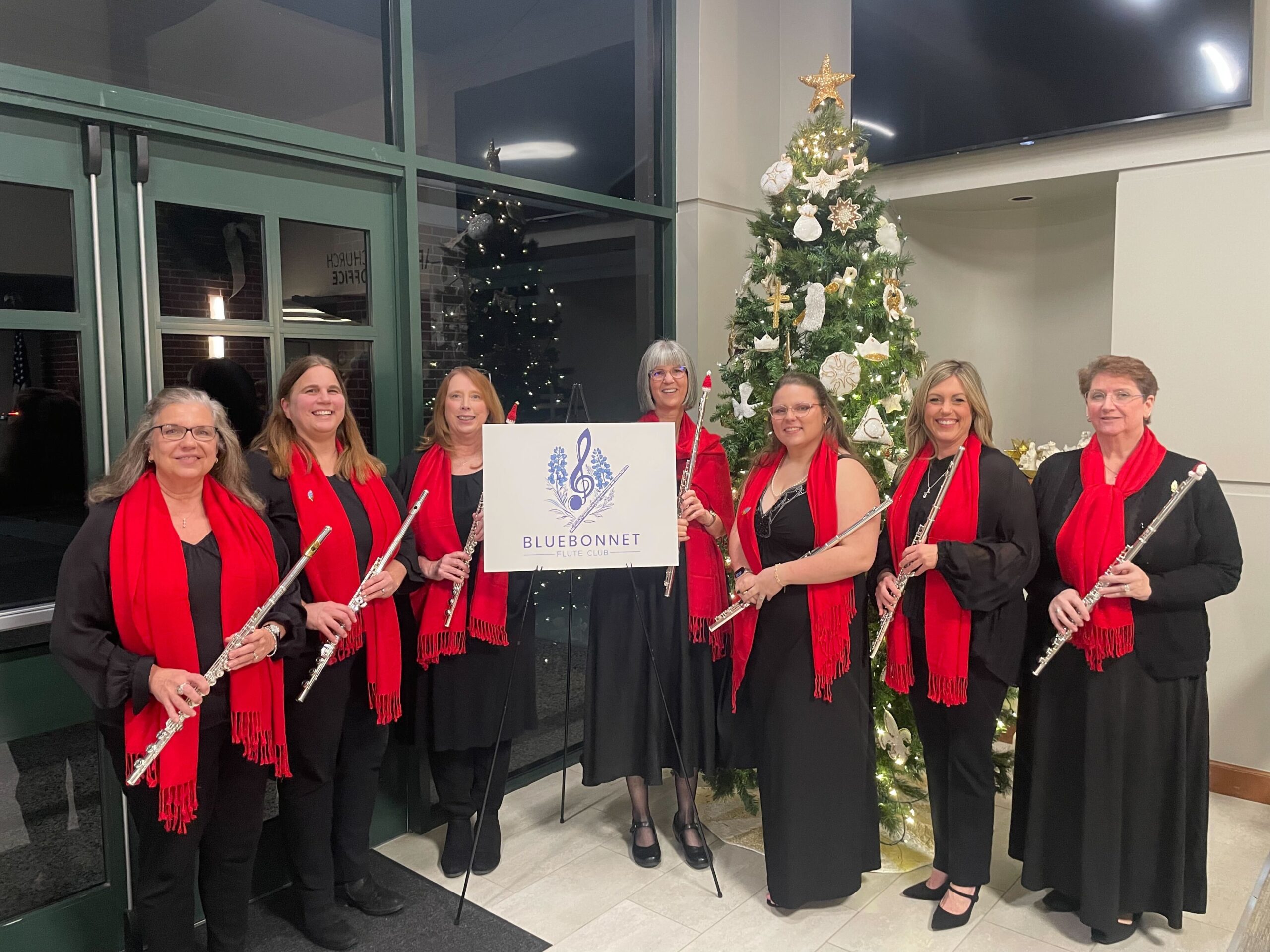 Group of women in black outfits and red scarves. They are holding flutes, standing in front of a Christmas tree and around an easel with the Bluebonnet Flute Club logo on it